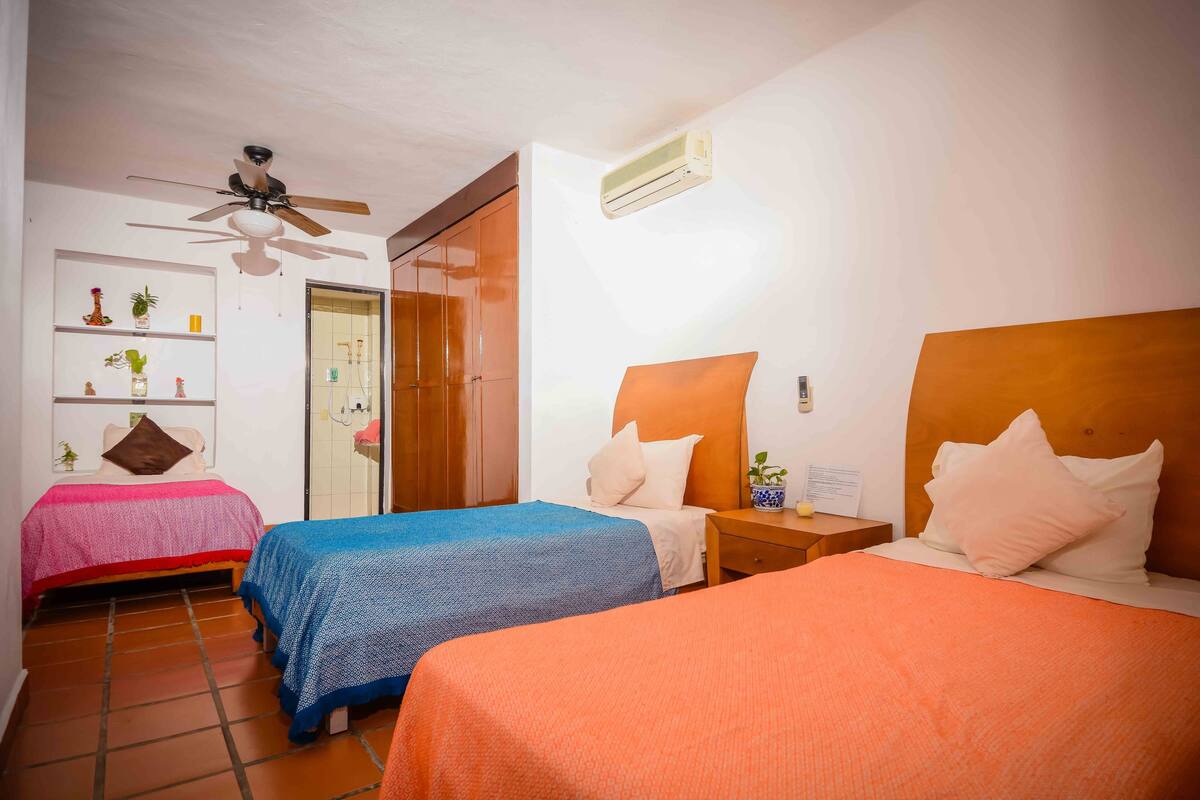Habitación de hotel con dos camas, ventilador de techo, aire acondicionado, mesita de noche y repisa con adornos.