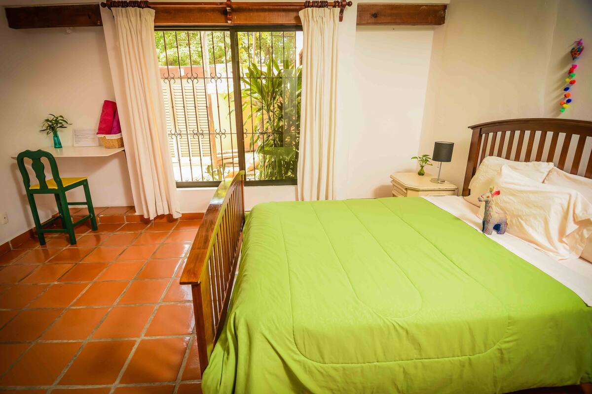 Una habitación con una cama, una silla, una mesita pequeña, una mesita de noche y una ventana con cortinas.