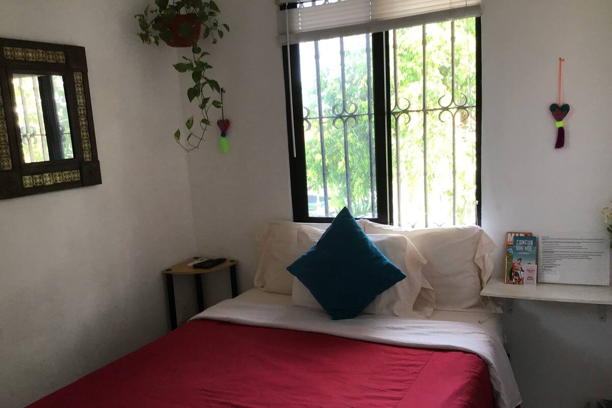 Una cama bien hecha con una colcha roja, una almohada azul y una almohada blanca sobre un colchón blanco. Se ve una mesita de noche pequeña con un control remoto encima. Se aprecia una ventana con barrotes, y una planta cuelga del techo.