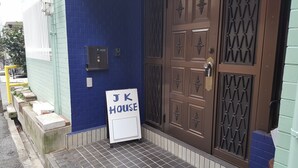 Une entrée d’immeuble avec un mur bleu, une porte marron ornée de panneaux décoratifs et une enseigne indiquant « JK HOUSE ».