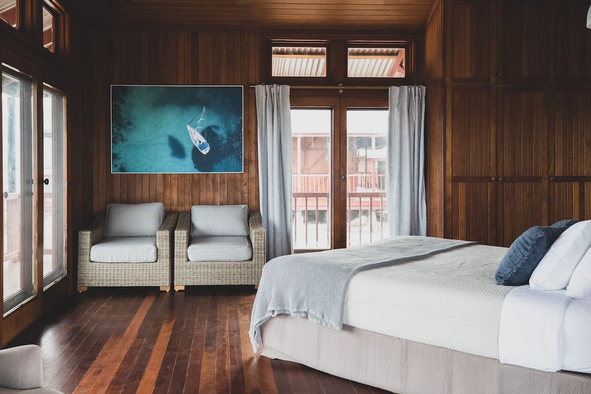 Quarto com uma cama grande, duas cadeiras de vime, teto de madeira e uma janela ampla com cortinas.