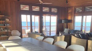 A spacious room with a large dining table, white chairs, a sofa with cushions, a bookshelf, and a view of the sea through large windows. - Camp Island Lodge (Camp Island)