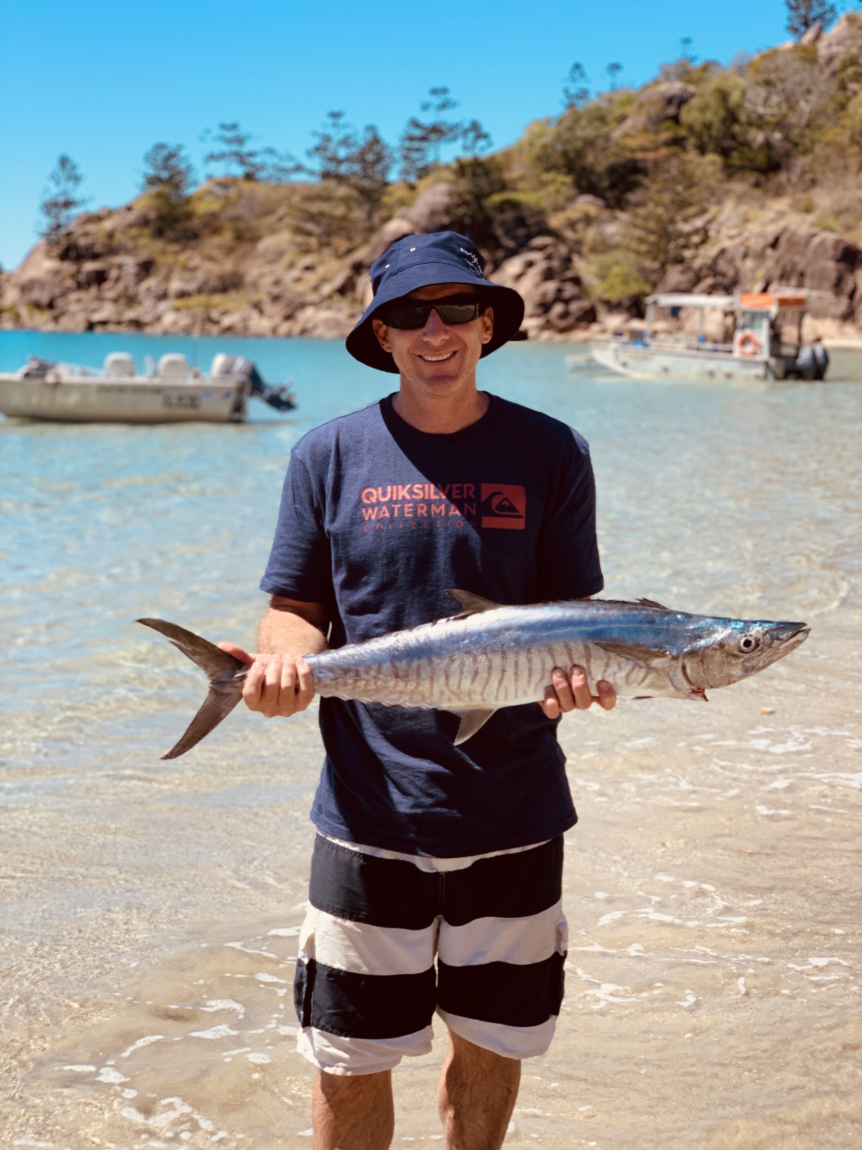 A person holding a fish on a beach with clear water and boats in the background.