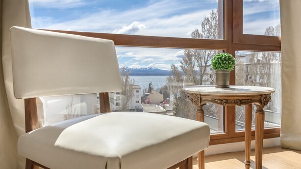 A room with a white chair, a small table with a plant, and a view of a lake and mountains through a large window. - DEPTO CENTRO C/PILETA Y VISTA AL LAGO N.HUAPI (San Carlos de Bariloche)