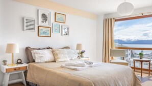A bedroom with a bed, bedside tables, lamps, a view of the mountains, and framed artwork on the wall. - DEPTO CENTRO C/PILETA Y VISTA AL LAGO N.HUAPI (San Carlos de Bariloche)