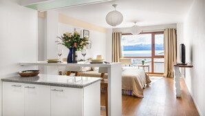A modern hotel room with a kitchen area, a dining table, a bed, and a large window offering a mountain view. - DEPTO CENTRO C/PILETA Y VISTA AL LAGO N.HUAPI (San Carlos de Bariloche)