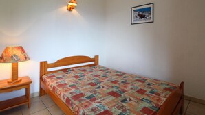 Un lit simple avec une couverture colorée, une table de chevet avec une lampe, un luminaire fixé au mur et un tableau encadré accroché au mur.