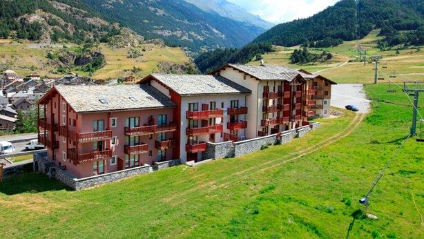 Un hôtel à plusieurs étages, doté de balcons, situé dans une région montagneuse, avec un télésiège visible au loin.