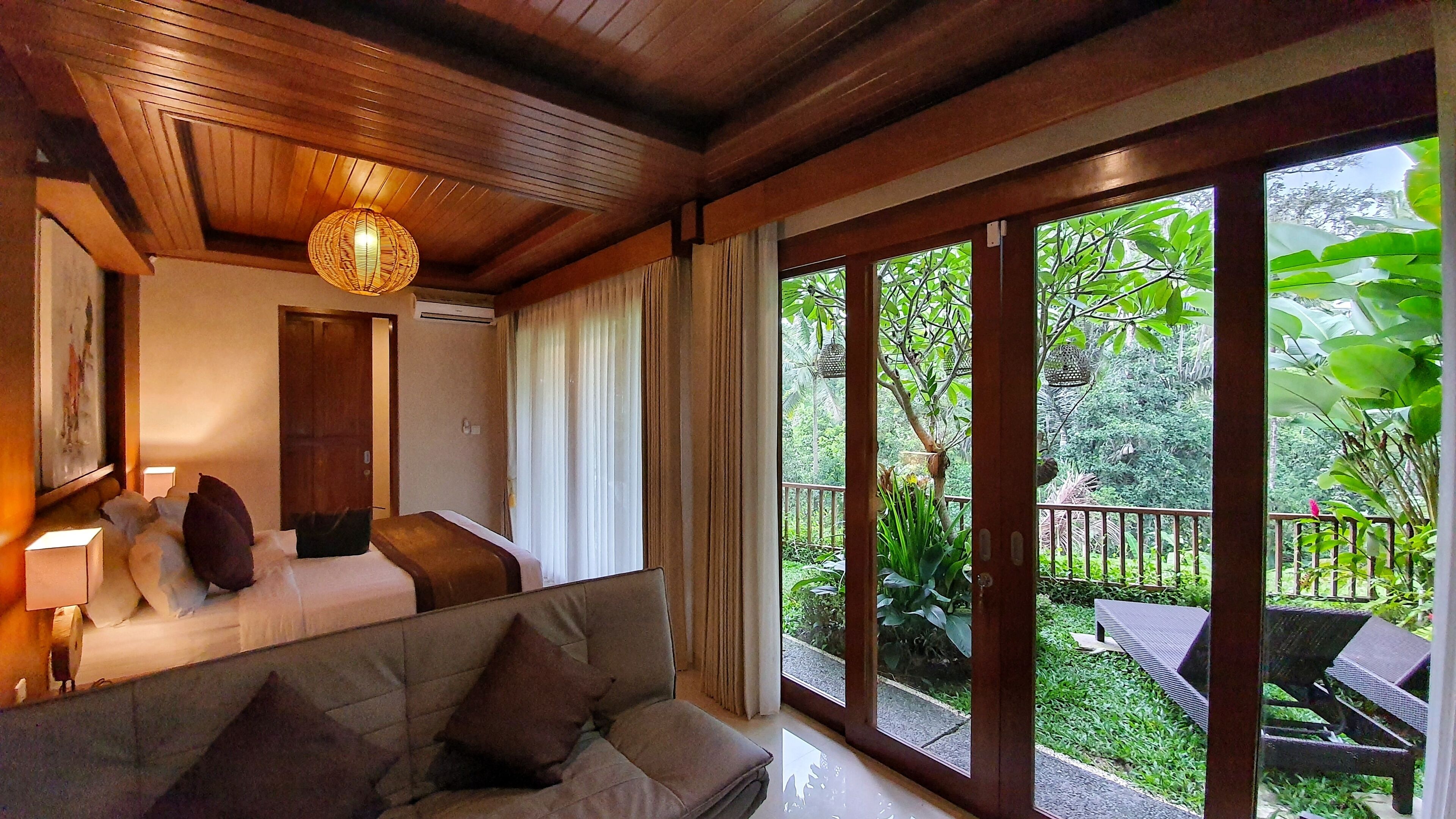 A room with a bed, a sofa, and a large window with a view of a garden.
