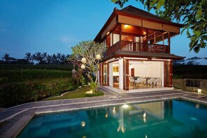 A two-story wooden house with a balcony, a swimming pool, and a dining area with outdoor furniture. - Puri Raya Villa Ubud (Tegallalang)