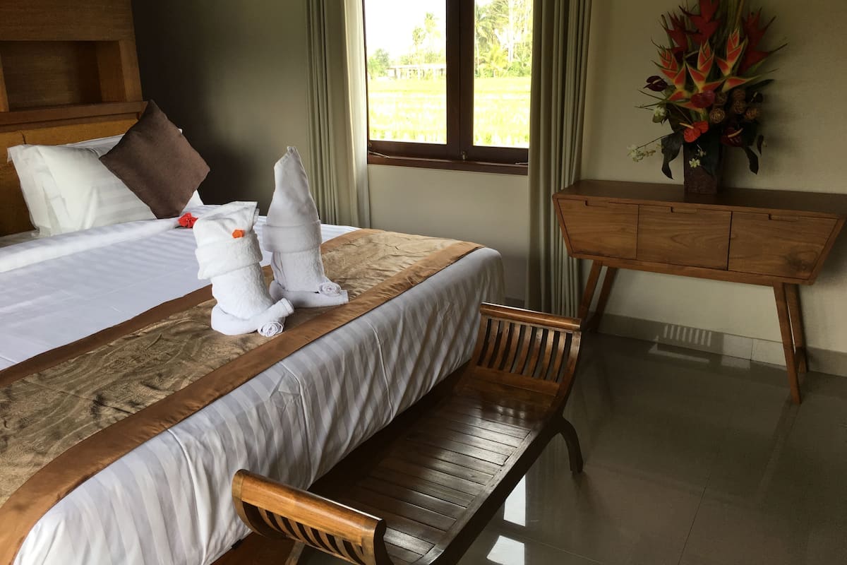 A hotel room with a bed, a wooden bench, a desk, and a window with a view of greenery.