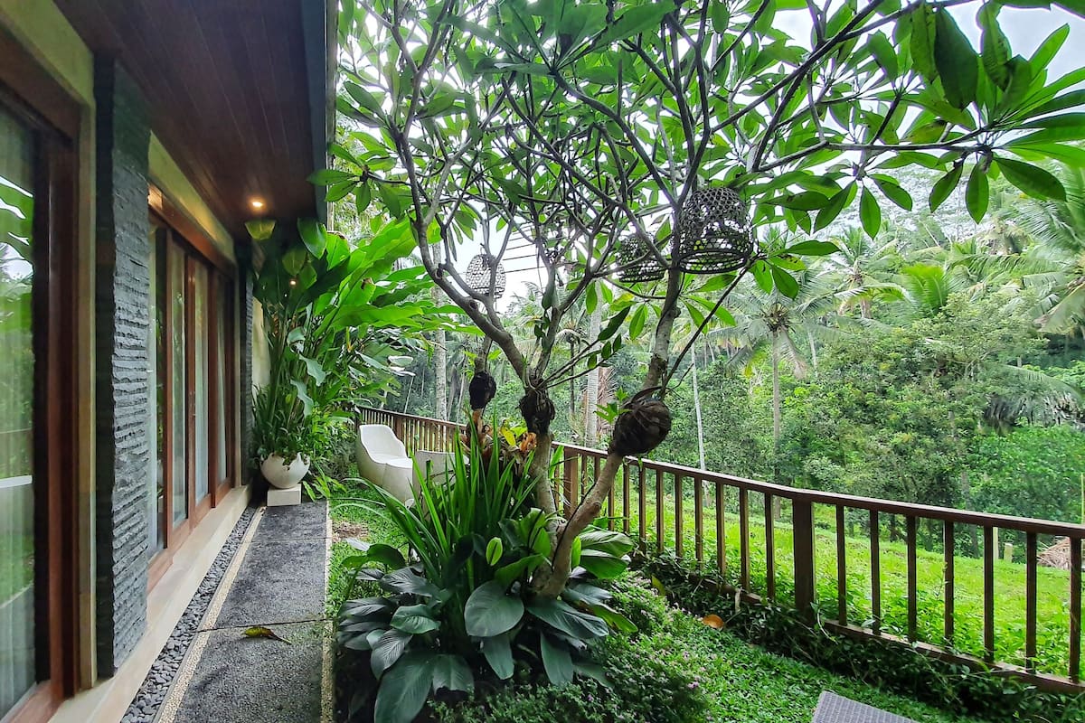 A modern building with large windows, a balcony with a railing, and lush greenery in the background.