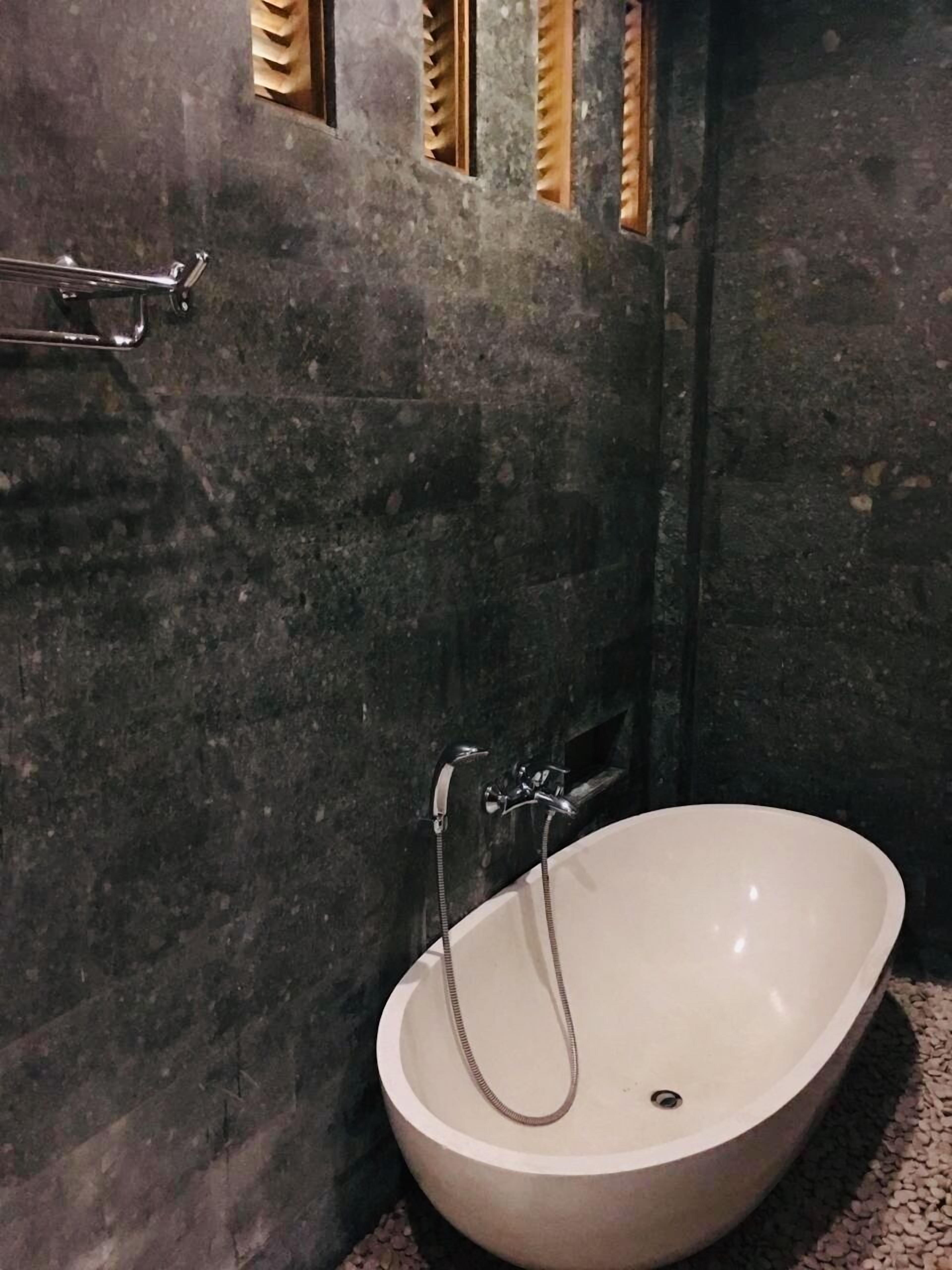 A bathroom with a freestanding bathtub, dark stone walls, and a small window with wooden blinds.