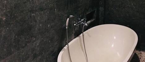 A bathroom with a freestanding bathtub, dark stone walls, and a small window with wooden blinds.