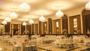 Un salón de banquetes con varias mesas redondas, sillas blancas y candelabros ornamentales.