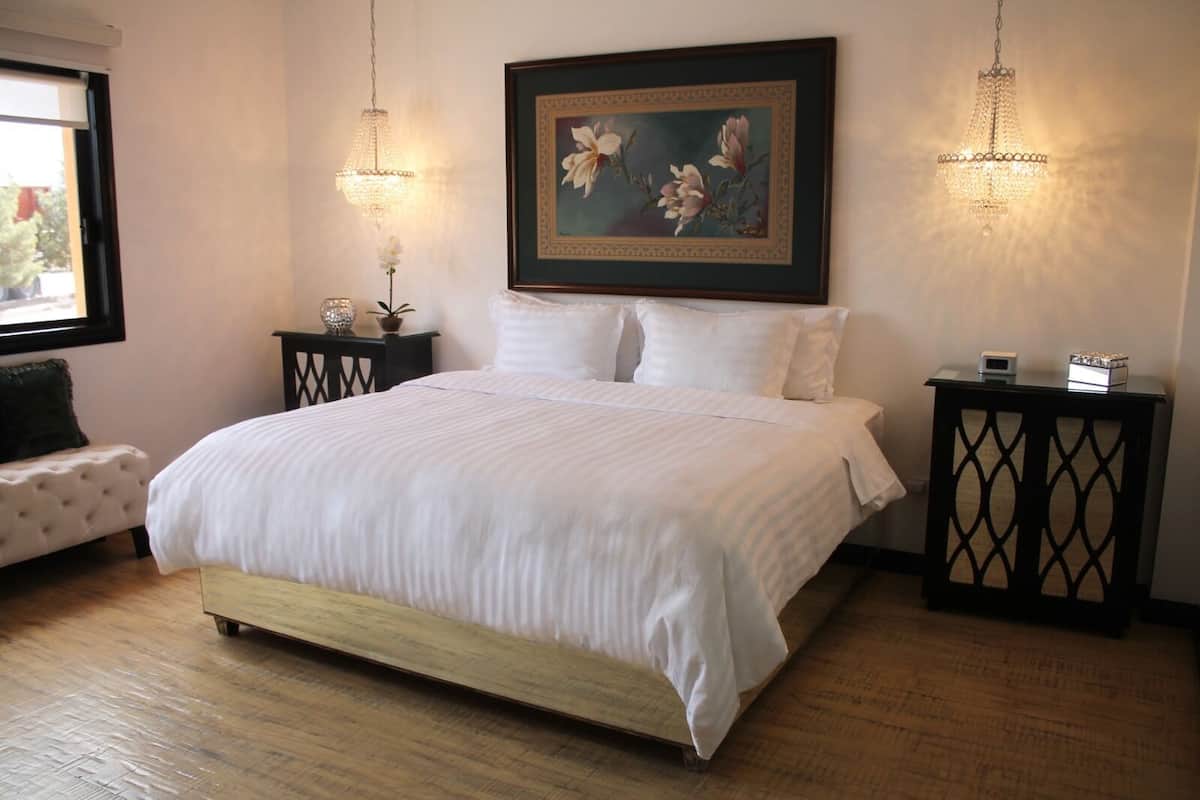 Un lit bien fait, avec du linge de lit blanc, une tête de lit en bois et un grand tableau floral au-dessus. On y trouve deux tables de chevet avec des lampes décoratives, une petite plante en pot et un canapé blanc dans le coin.