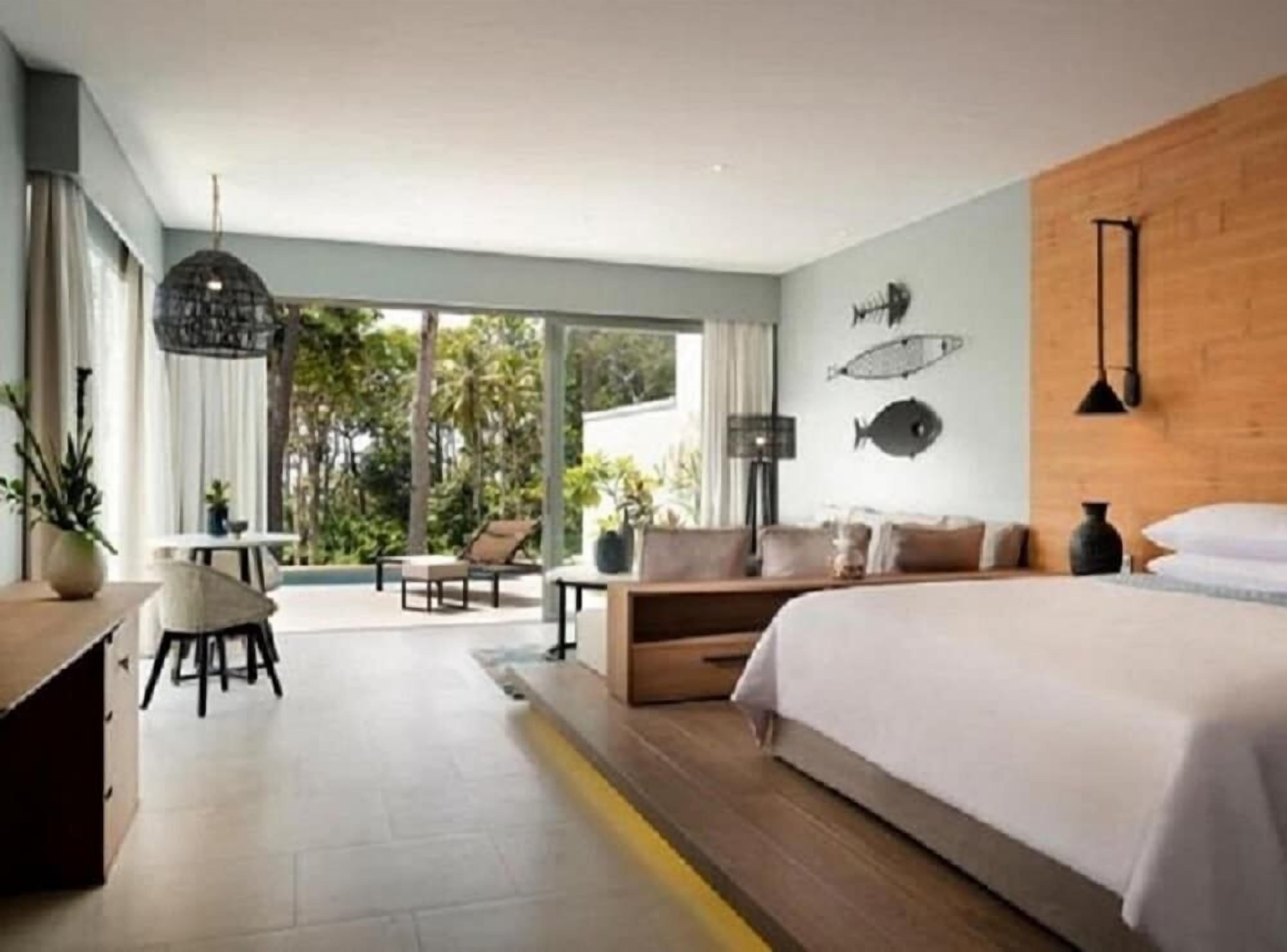 A modern hotel room with a large bed, a sofa, a dining area with a table and chairs, and a balcony with a view of greenery.