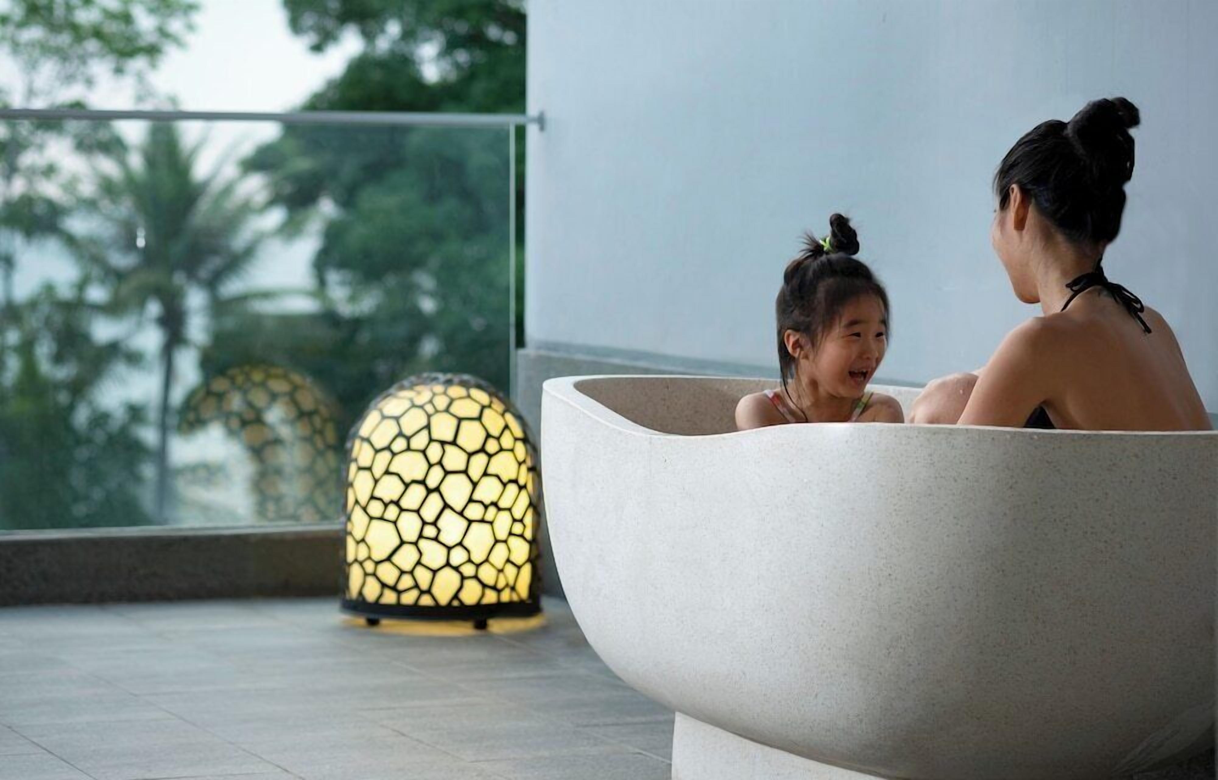 Two people sitting in a large, modern bathtub in a spacious room with a large window and a decorative lamp.