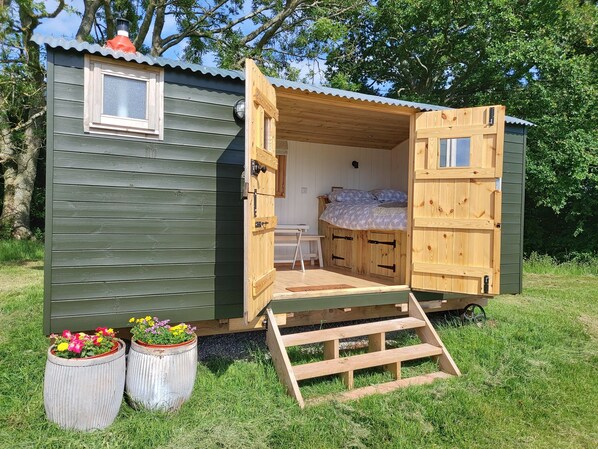 Una pequeña cabaña de madera con una cama, una ventana y una puerta. Hay dos macetas con flores afuera.