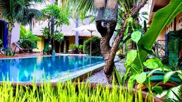 Un área de piscina con un árbol y jaulas colgantes para pájaros, rodeada de abundante follaje y una estructura con techo de paja.