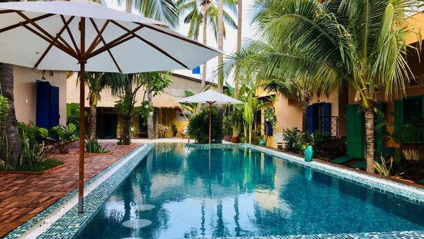A swimming pool with a tiled edge, surrounded by palm trees and a building with blue shutters. - MiNhon Hotel Mui Ne (Mui Ne)