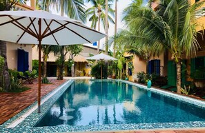 Una piscina con borde de azulejos, rodeada de palmeras y un edificio con persianas azules.