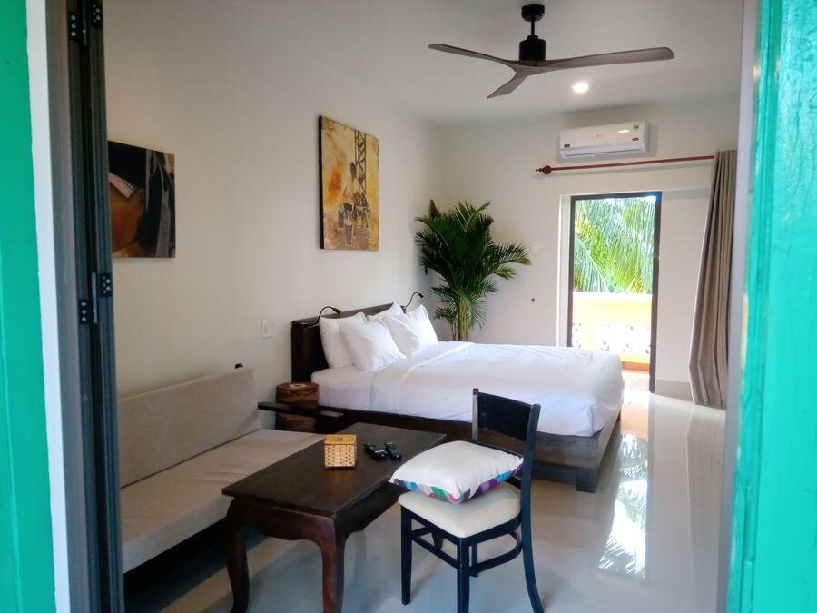 A hotel room with a bed, a sofa, a table, a chair, a plant, and an air conditioning unit.