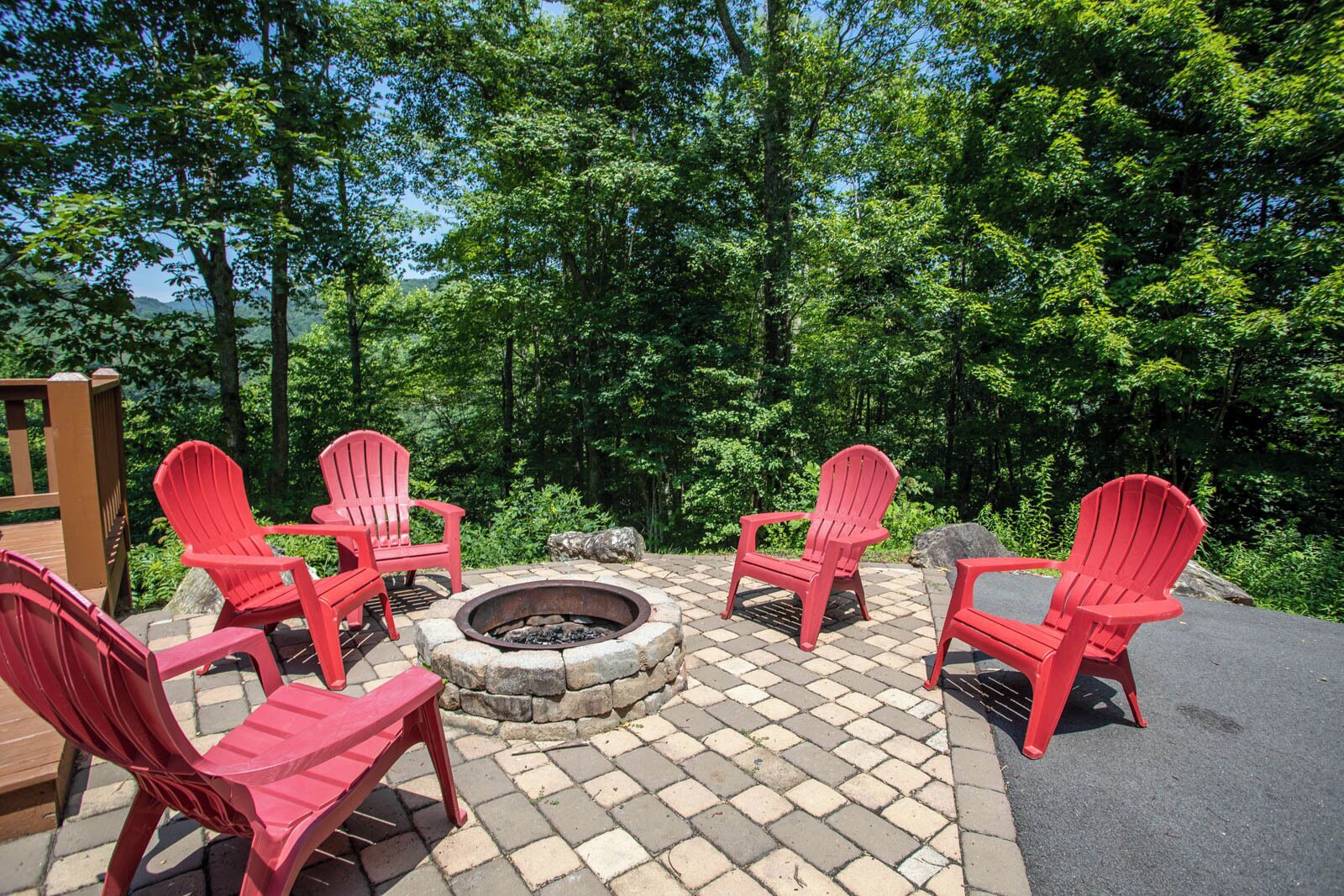 A fire pit surrounded by red Adirondack chairs in a forested outdoor area.