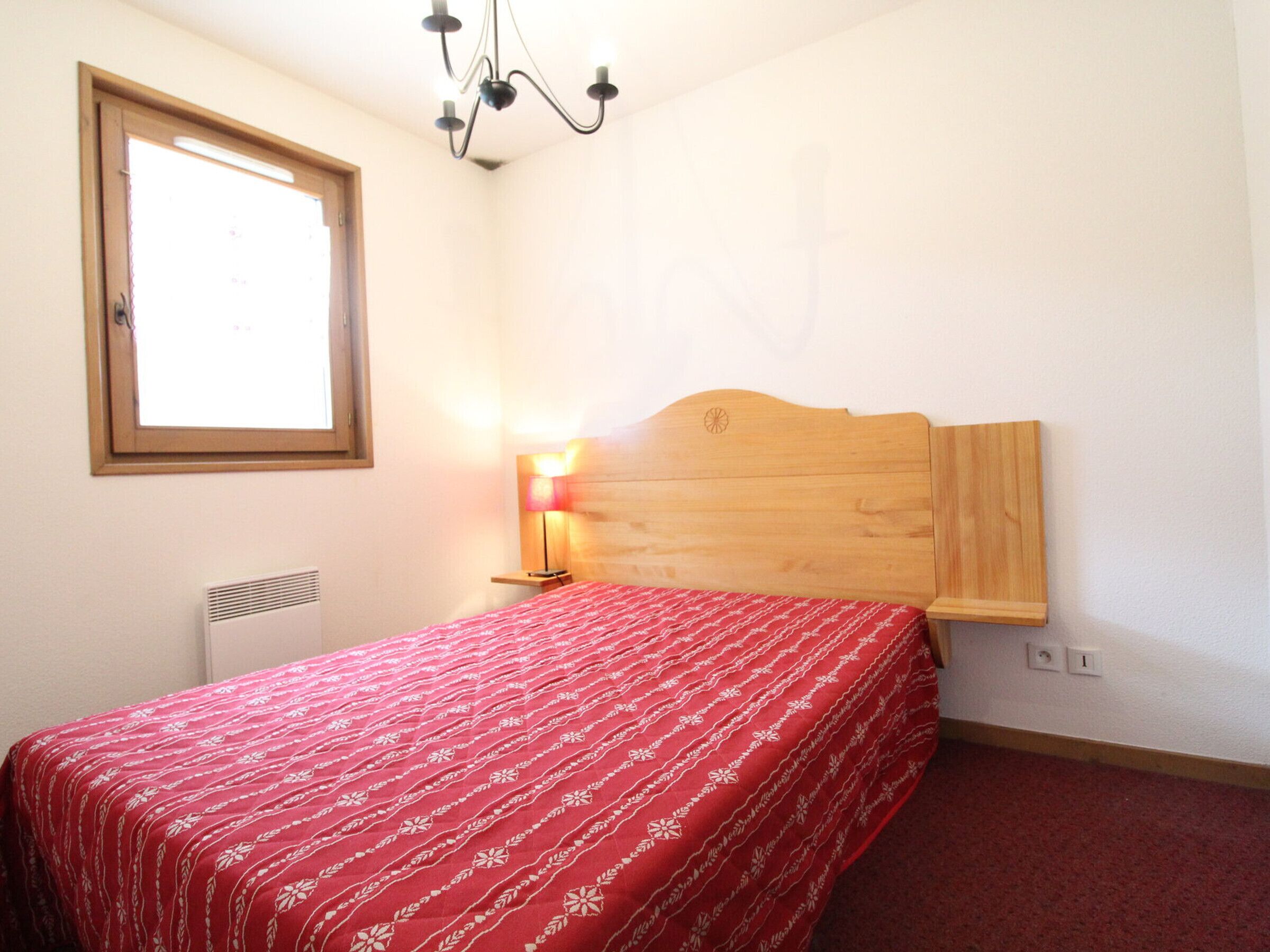 A bedroom with a wooden headboard, a red bedspread, a bedside lamp, a window, and a radiator.