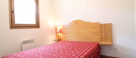 A bedroom with a wooden headboard, a red bedspread, a bedside lamp, a window, and a radiator.