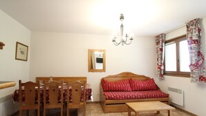 A room with a wooden dining table, chairs, a sofa with red cushions, a wooden coffee table, a chandelier, a window with patterned curtains, and a mirror on the wall. - Spacious apartment for 6 people at the foot of the slopes with parking and WIFI (Lanslevillard)