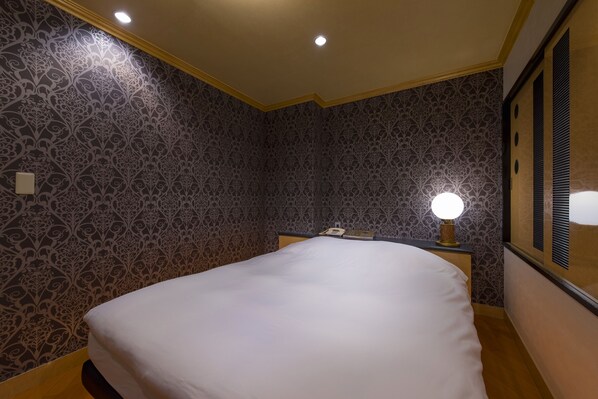 A hotel room with a large bed, patterned wallpaper, a bedside table with a lamp, and a phone. - Hotel Linden Oji (Tokyo)