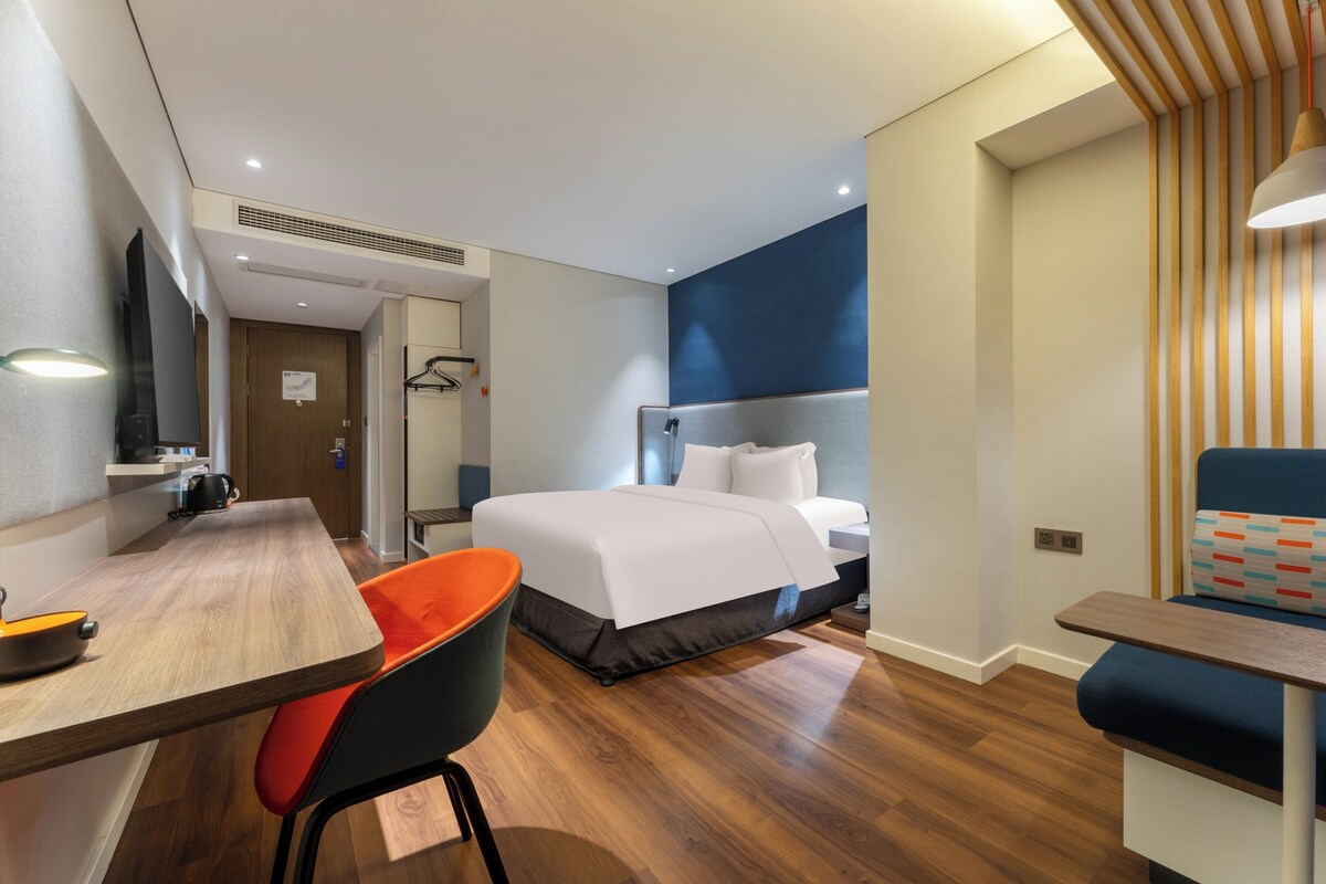 A modern hotel room with a wooden floor, a bed with white bedding, a desk with a chair, and a TV.