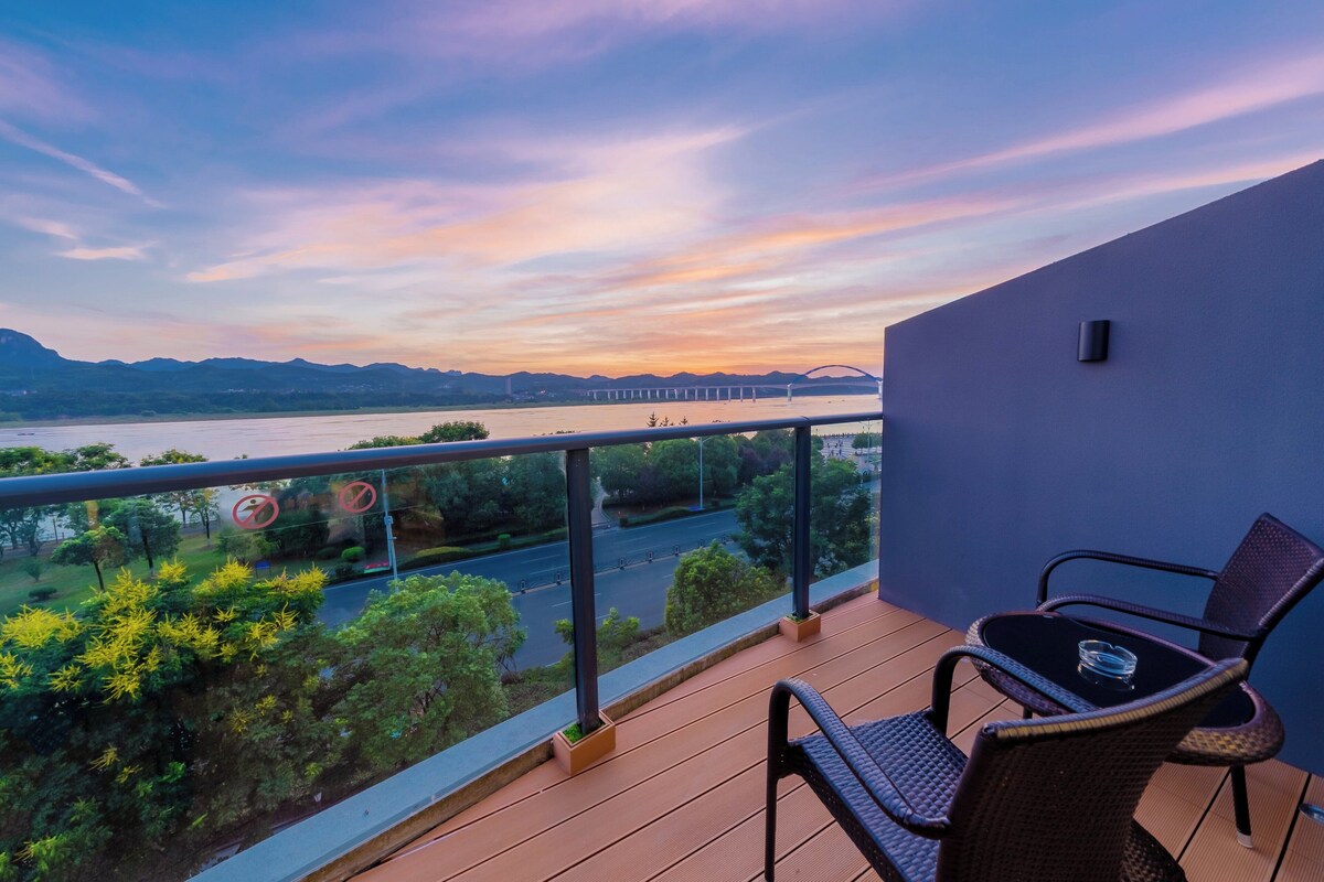A balcony with a view of a river and mountains, a table with a chair, and a railing.