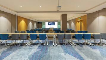 Un salón de conferencias con una larga mesa, filas de sillas y una pantalla de proyección.