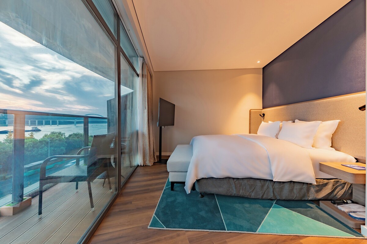 A modern hotel room with a large bed, a view of the cityscape, and a balcony with a chair.