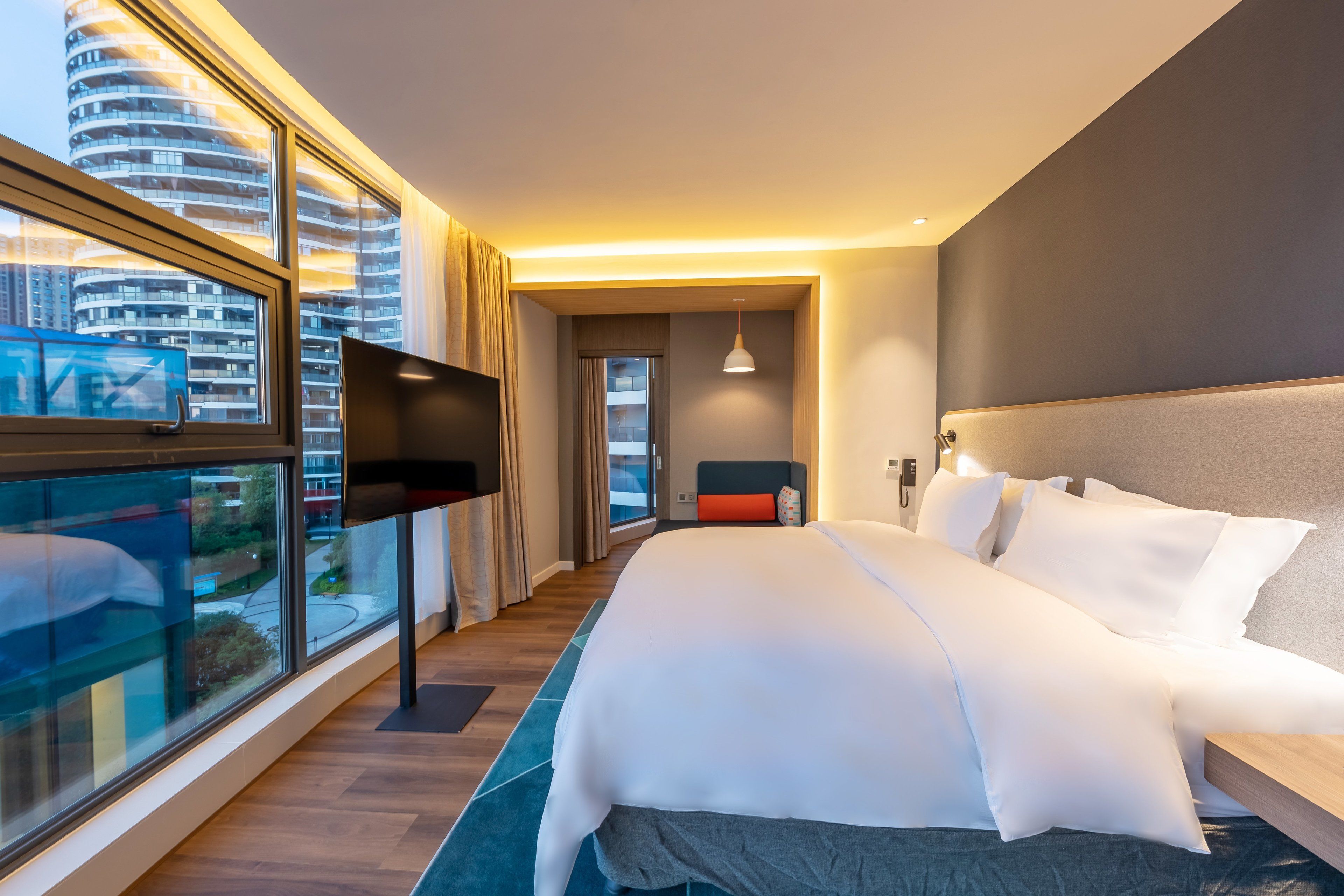 A modern hotel room with a large bed, a TV, and a view of the cityscape through large windows.