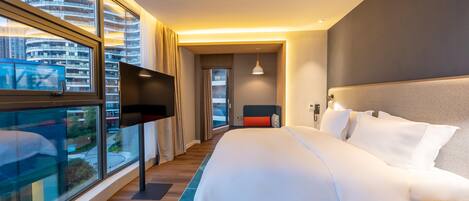 A modern hotel room with a large bed, a TV, and a view of the cityscape through large windows.