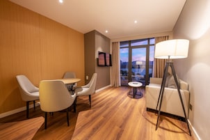 Una habitación de hotel moderna con piso de madera, un sofá, dos sillones, una mesita redonda, un televisor y una lámpara de pie.