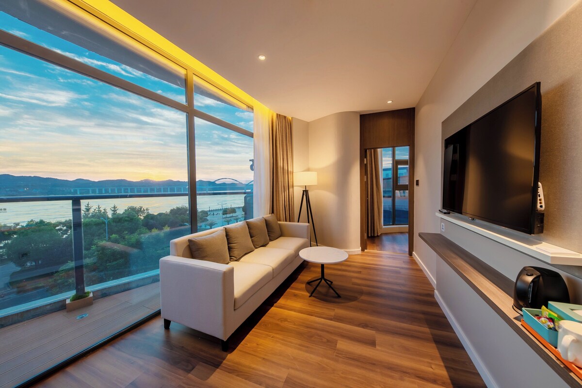 A modern living room with a sofa, a small round table, a TV, and a view of a bridge over water.