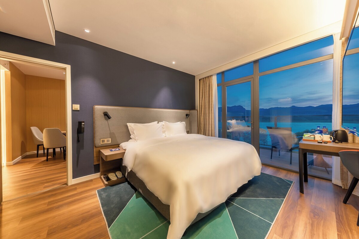 A hotel room with a large bed, a desk with a chair, a phone, and a view of the mountains through the window.