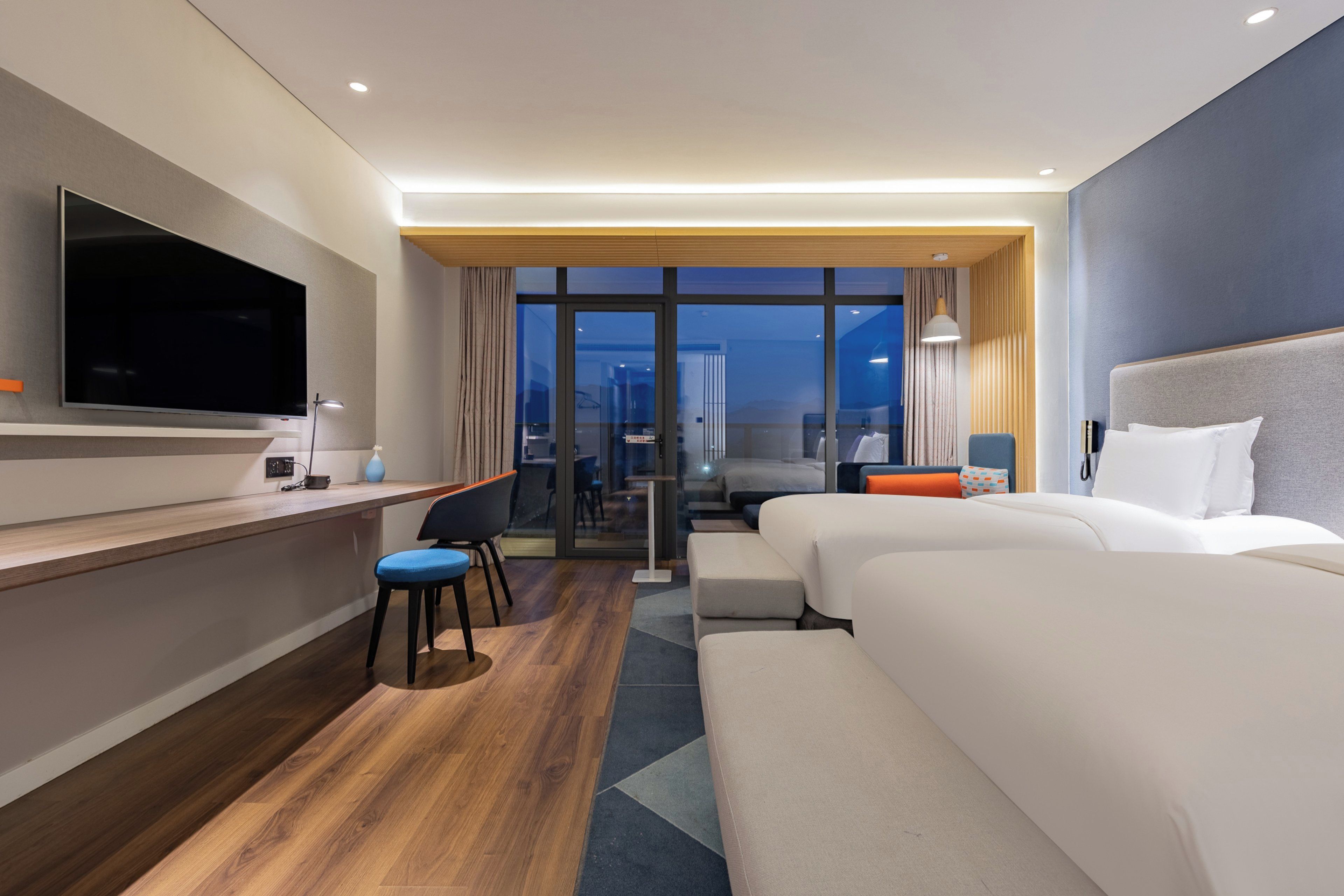 Une chambre d’hôtel moderne équipée d’une télévision à écran plat, d’un bureau avec une lampe, de deux lits, d’un canapé et offrant une vue sur l’extérieur grâce à de grandes fenêtres.