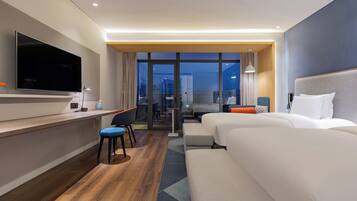 Une chambre d’hôtel moderne équipée d’une télévision à écran plat, d’un bureau avec une lampe, de deux lits, d’un canapé et offrant une vue sur l’extérieur grâce à de grandes fenêtres.