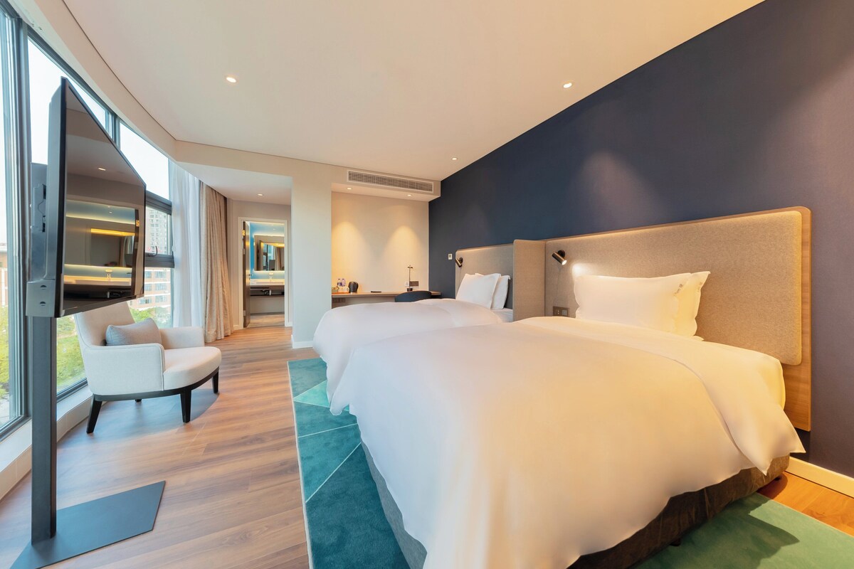 A modern hotel room with a large bed, a seating area, and a view through the window.