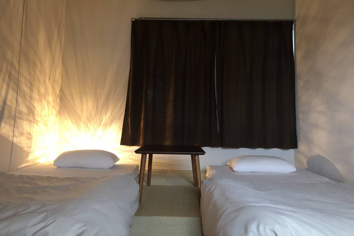 Quarto aconchegante com duas camas, uma mesinha e uma janela com cortinas.