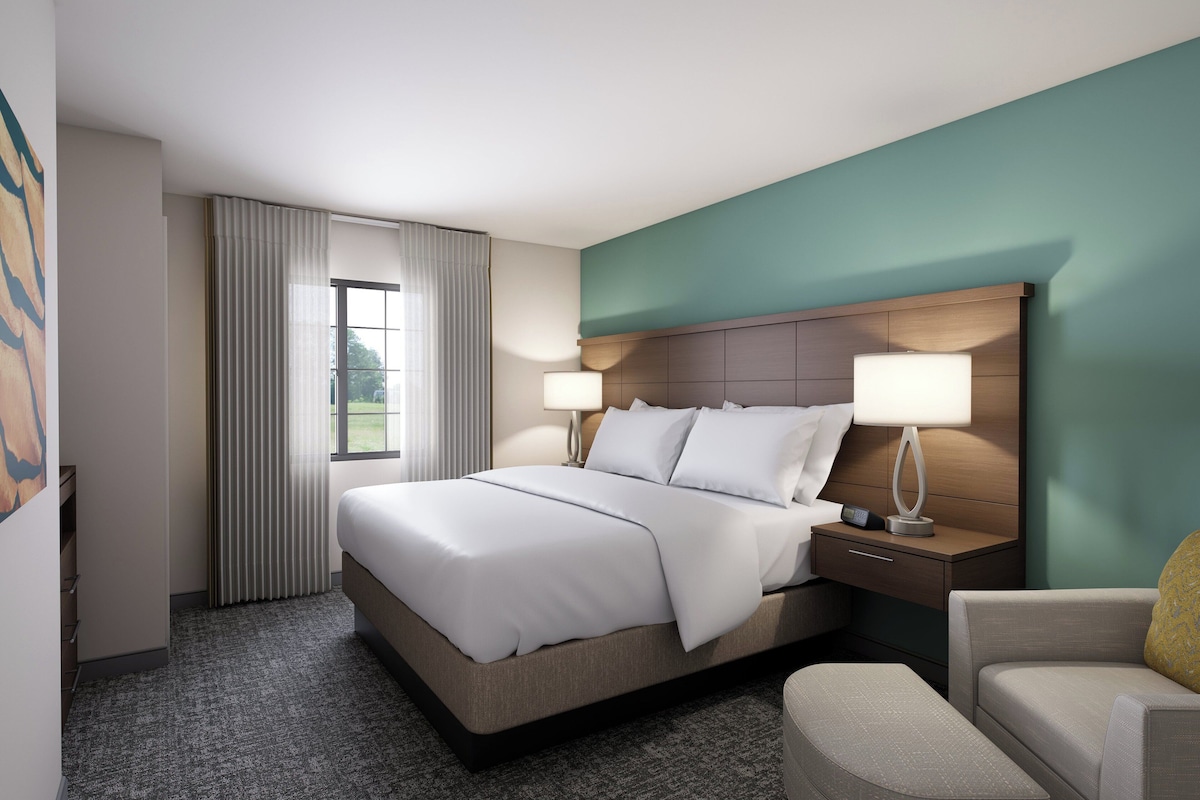 A hotel room with a large bed, bedside lamps, a nightstand, a chair, and a window with curtains.