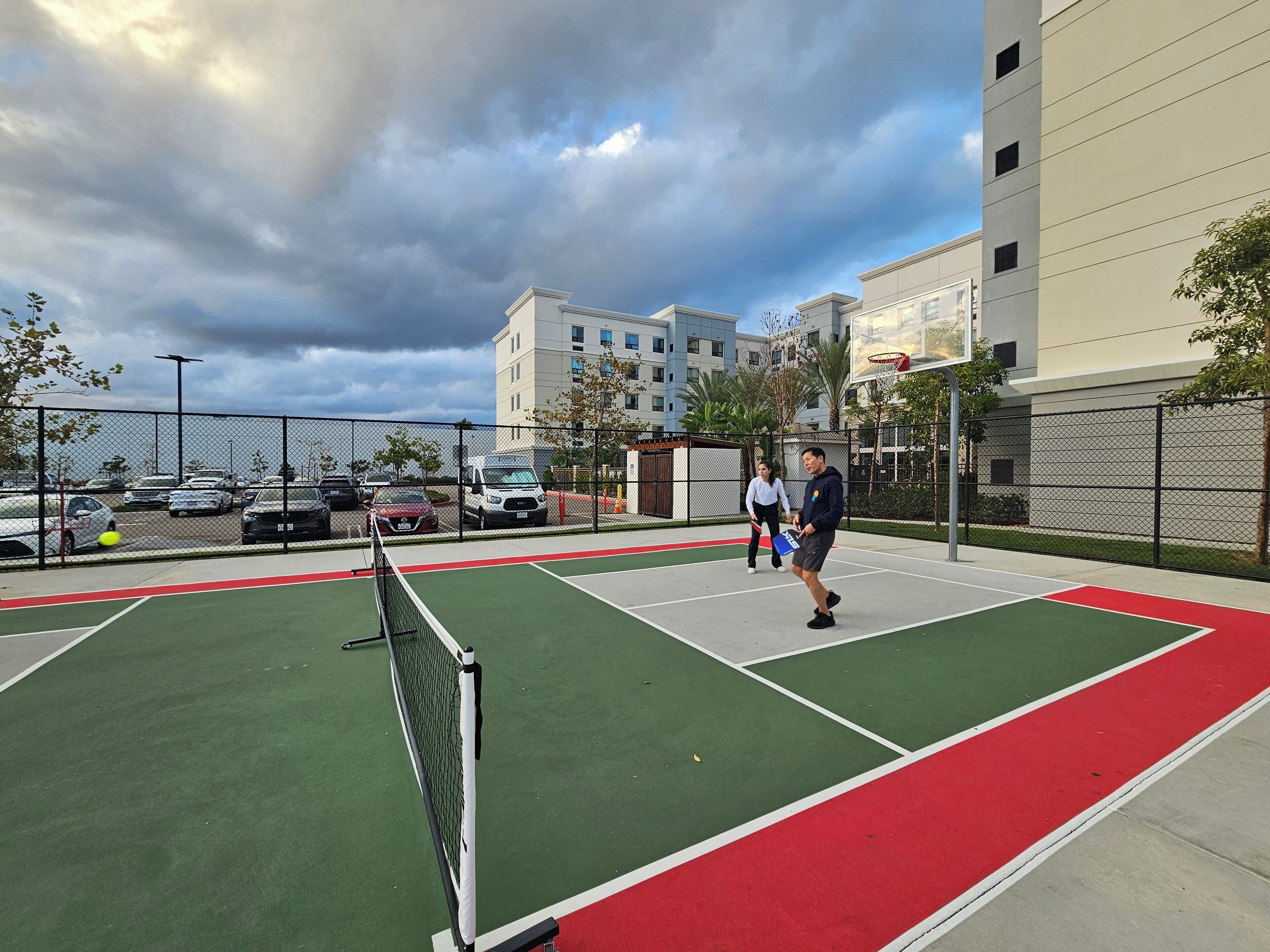 Cancha de tenis al aire libre con red, rodeada por una valla y contigua a un estacionamiento con automóviles.