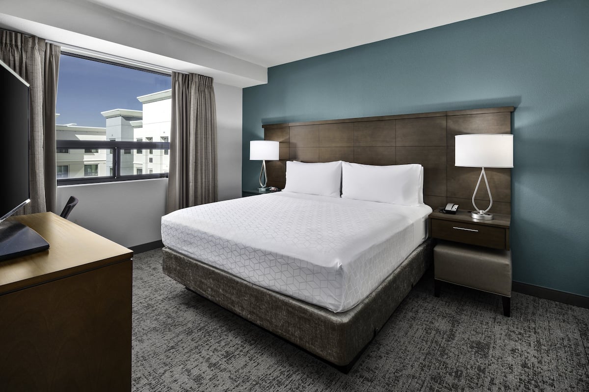 A hotel room with a large bed, two bedside tables with lamps, a desk, and a view of buildings through the window.