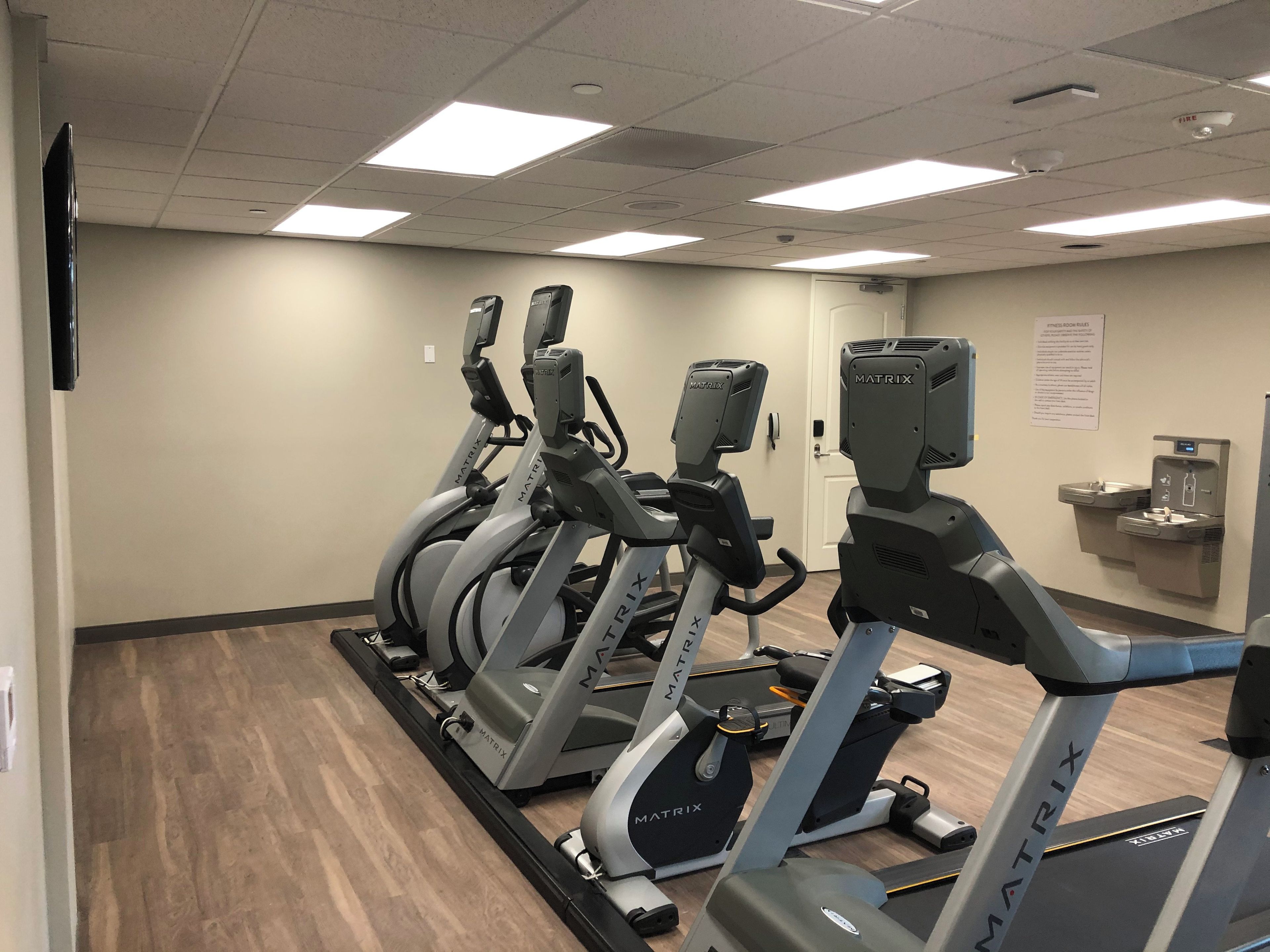 Varias bicicletas estáticas alineadas en un gimnasio con suelo de madera, pared blanca y una puerta al fondo.