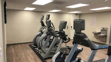 Varias bicicletas estáticas alineadas en un gimnasio con suelo de madera, pared blanca y una puerta al fondo.